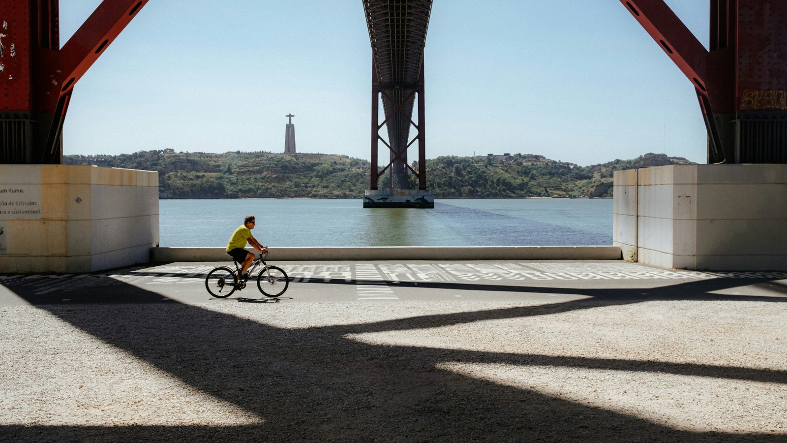 A cyclist riding along the river beneath the red steel of a suspension bridge